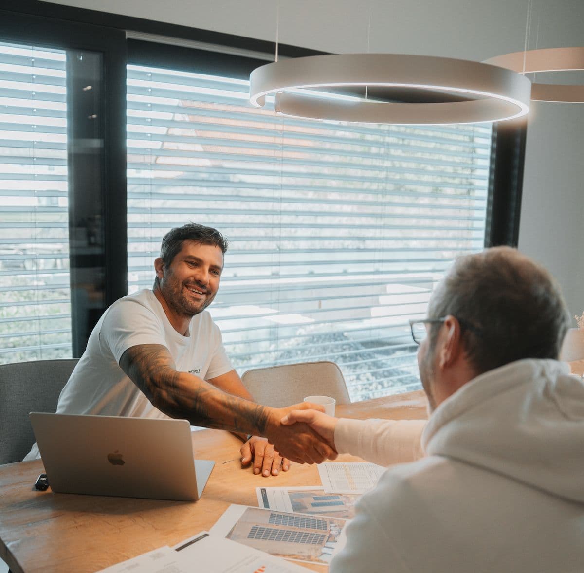 Freundlicher Handschlag zwischen zwei Männern am Tisch mit Arbeitsunterlagen zur erfolgreichen Partnerschaft bei Droneworks.