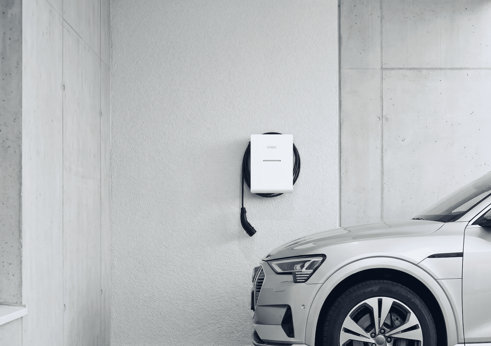 Weiße SENEC Ladestation an einer strukturierten Wand in einer modernen Garage aus Sichtbeton mit einem silbernen E-Auto.